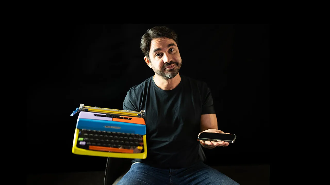 Marcelo Albuquerque sentado, segurando uma máquina de escrever antiga e um smartphone